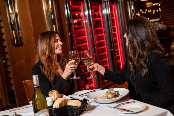 A restaurant interior with a wine rack in the background, a bottle of wine, and plates of food on the table. - Lady Luck Casino Black Hawk - A Caesars Rewards Destination (Black Hawk)