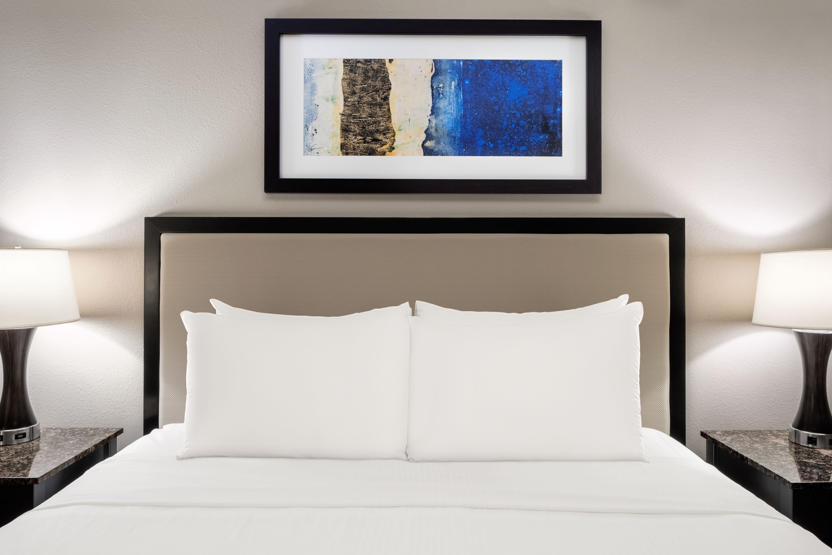 A neatly made bed with white linens and pillows, a framed artwork above it, and two bedside lamps on either side.