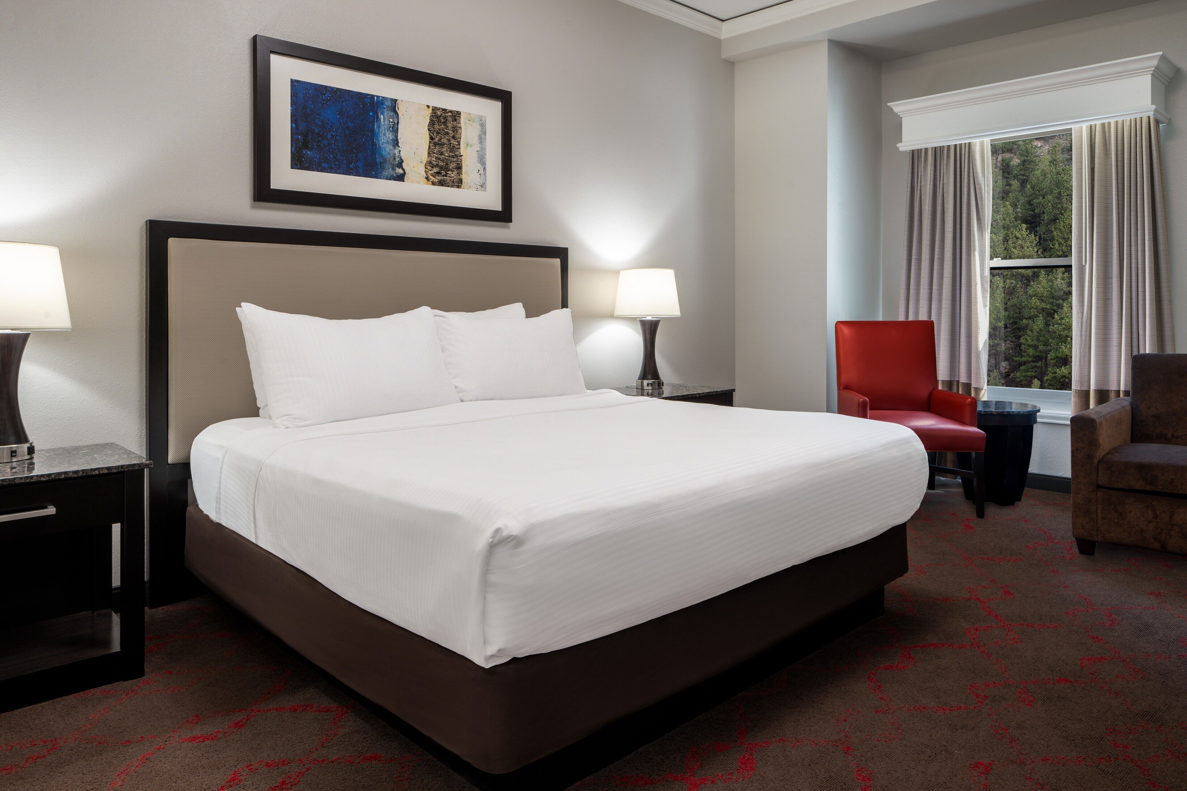 A hotel room with a large bed, two bedside lamps, a red chair, a window with curtains, and a framed artwork on the wall.