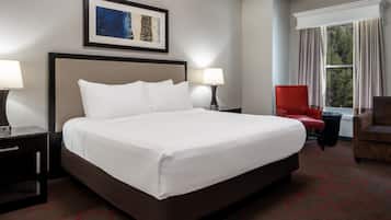 A hotel room with a large bed, two bedside lamps, a red chair, a window with curtains, and a framed artwork on the wall.