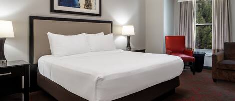 A hotel room with a large bed, two bedside lamps, a red chair, a window with curtains, and a framed artwork on the wall.