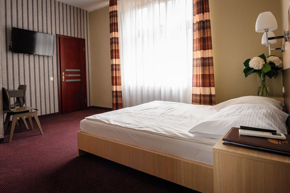 A hotel room with a bed, a desk, a chair, a TV mounted on the wall, and a window with curtains.
