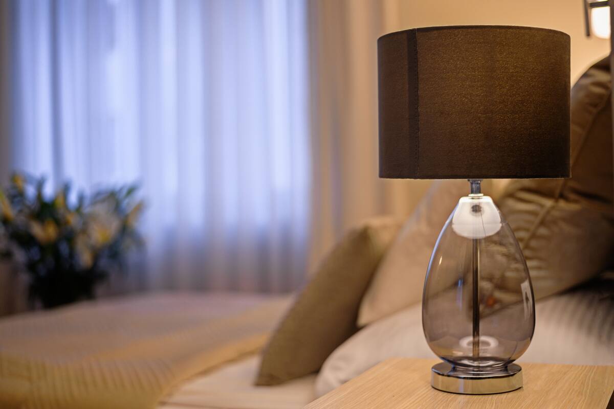 Une lampe moderne, dotée d’un pied transparent et d’un abat-jour foncé, posée sur une table d’appoint en bois dans un salon.