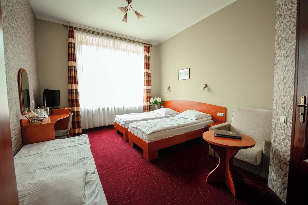Une chambre d’hôtel avec deux lits, un bureau, une chaise, une petite table, un téléviseur et une fenêtre avec des rideaux.
