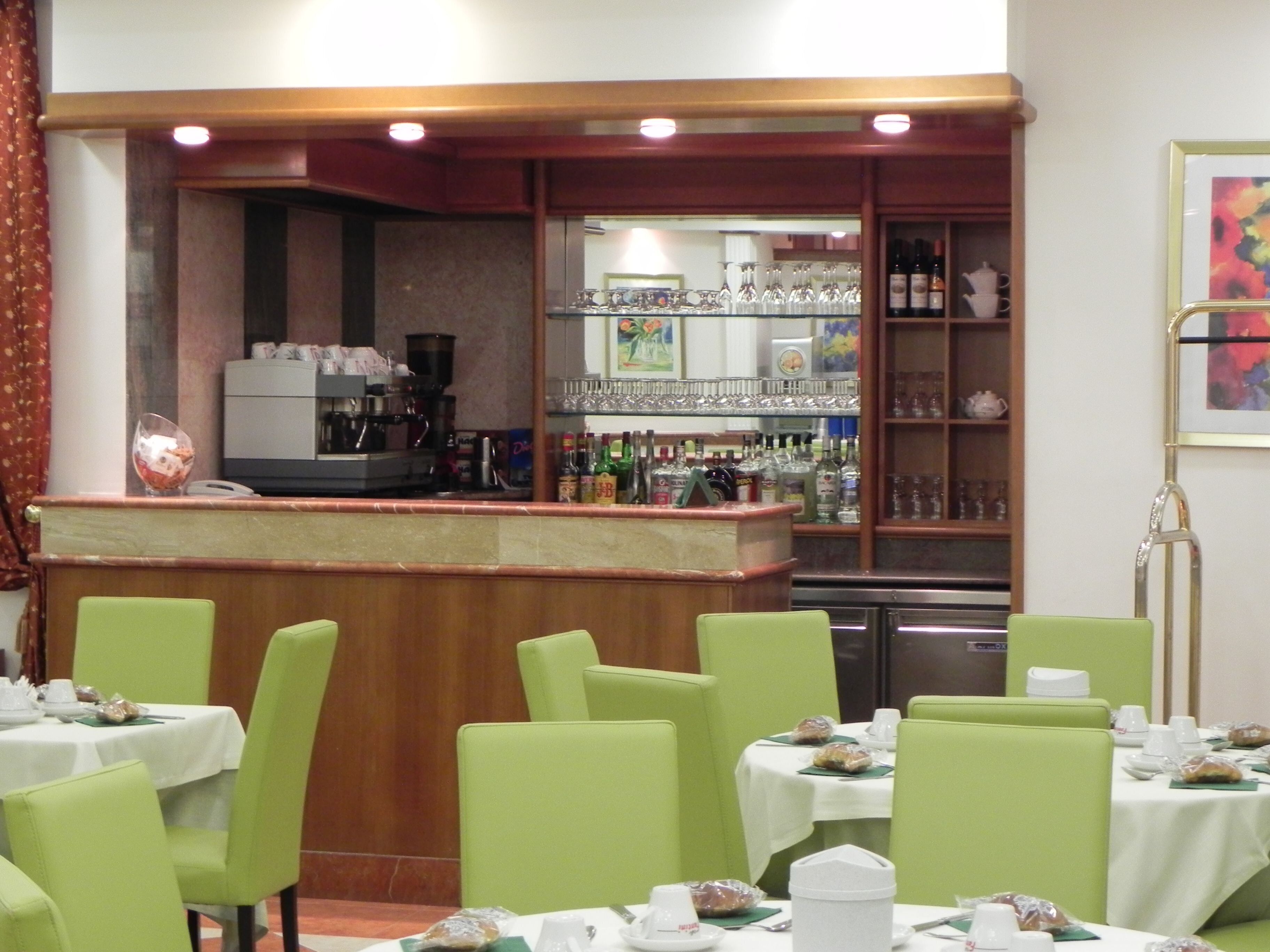 A restaurant with green chairs and tables, a bar area with a counter, and shelves with bottles and glasses.