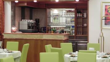 A restaurant with green chairs and tables, a bar area with a counter, and shelves with bottles and glasses.