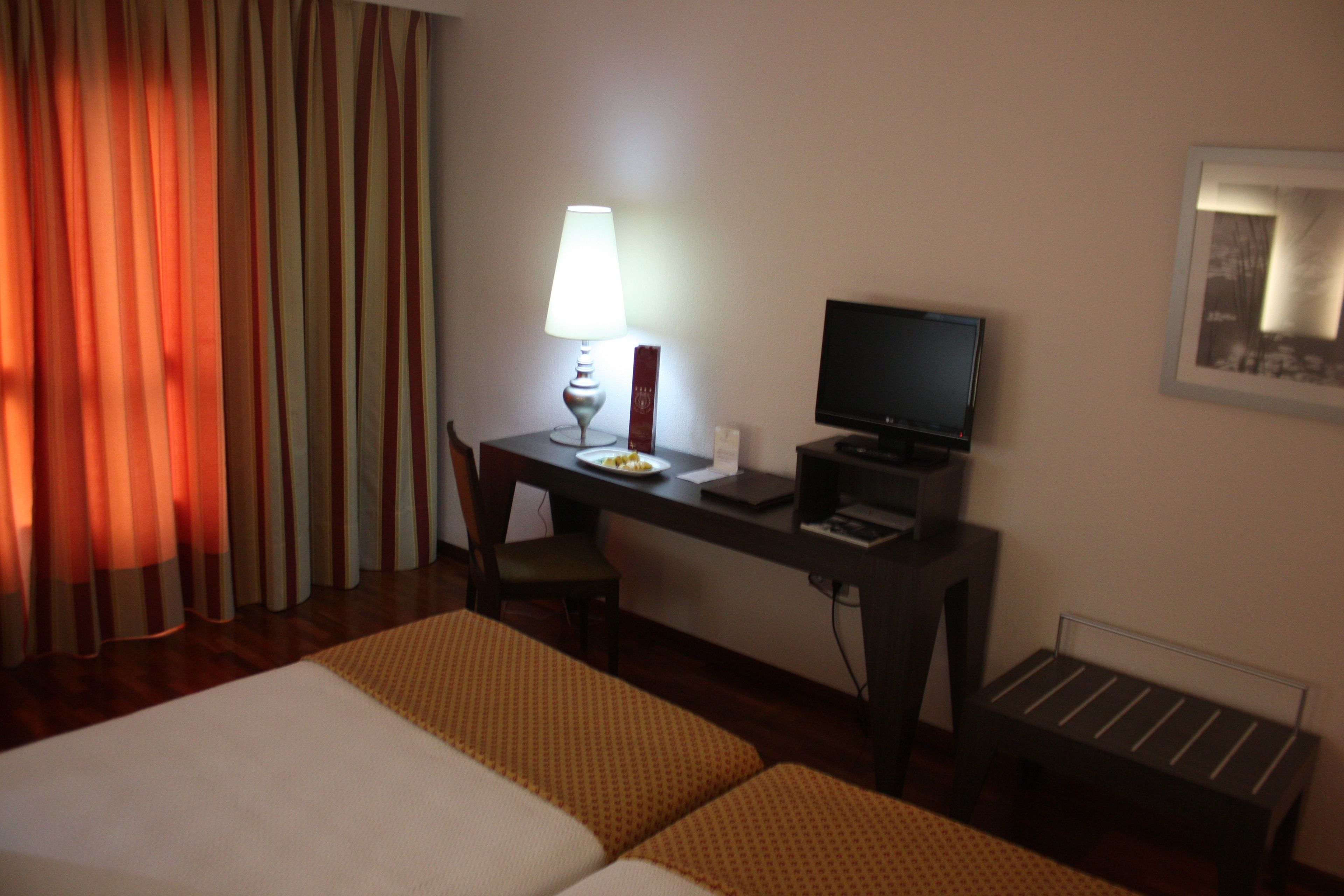 A hotel room with two beds, a desk with a computer, a lamp, and a small table.
