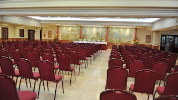 Un salón amplio con hileras de sillas rojas, una mesa larga con mantel blanco y un mural en la pared.