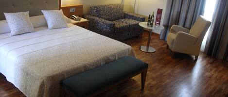 Habitación de hotel con una cama grande, un banco, una silla, un sofá, una mesita y una lámpara.