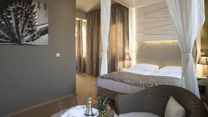Habitación de hotel con una cama, una silla de mimbre, una mesa con una botella y fruta, y un cuadro en la pared.
