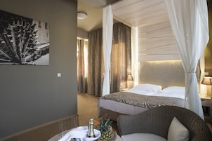 A hotel room with a bed, a wicker chair, a table with a bottle and fruit, and a painting on the wall.