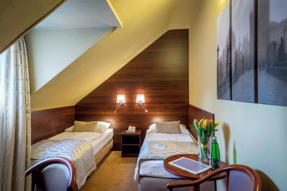 Une chambre d’hôtel avec deux lits simples, une petite table avec une chaise, une table de chevet avec un téléphone et une lampe fixée au mur.