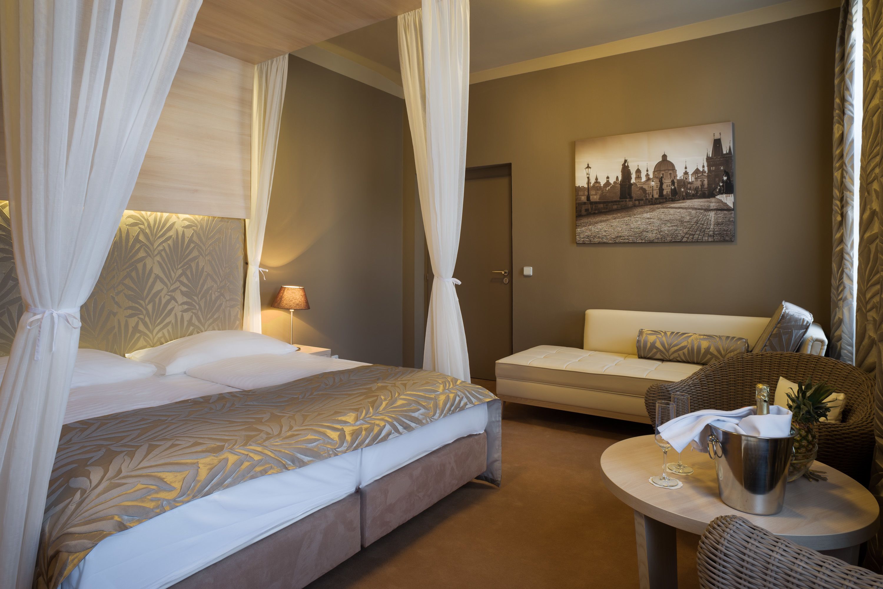 A hotel room with a bed, a sofa, a wicker chair, a small table with a drink and a pineapple, and a painting on the wall.