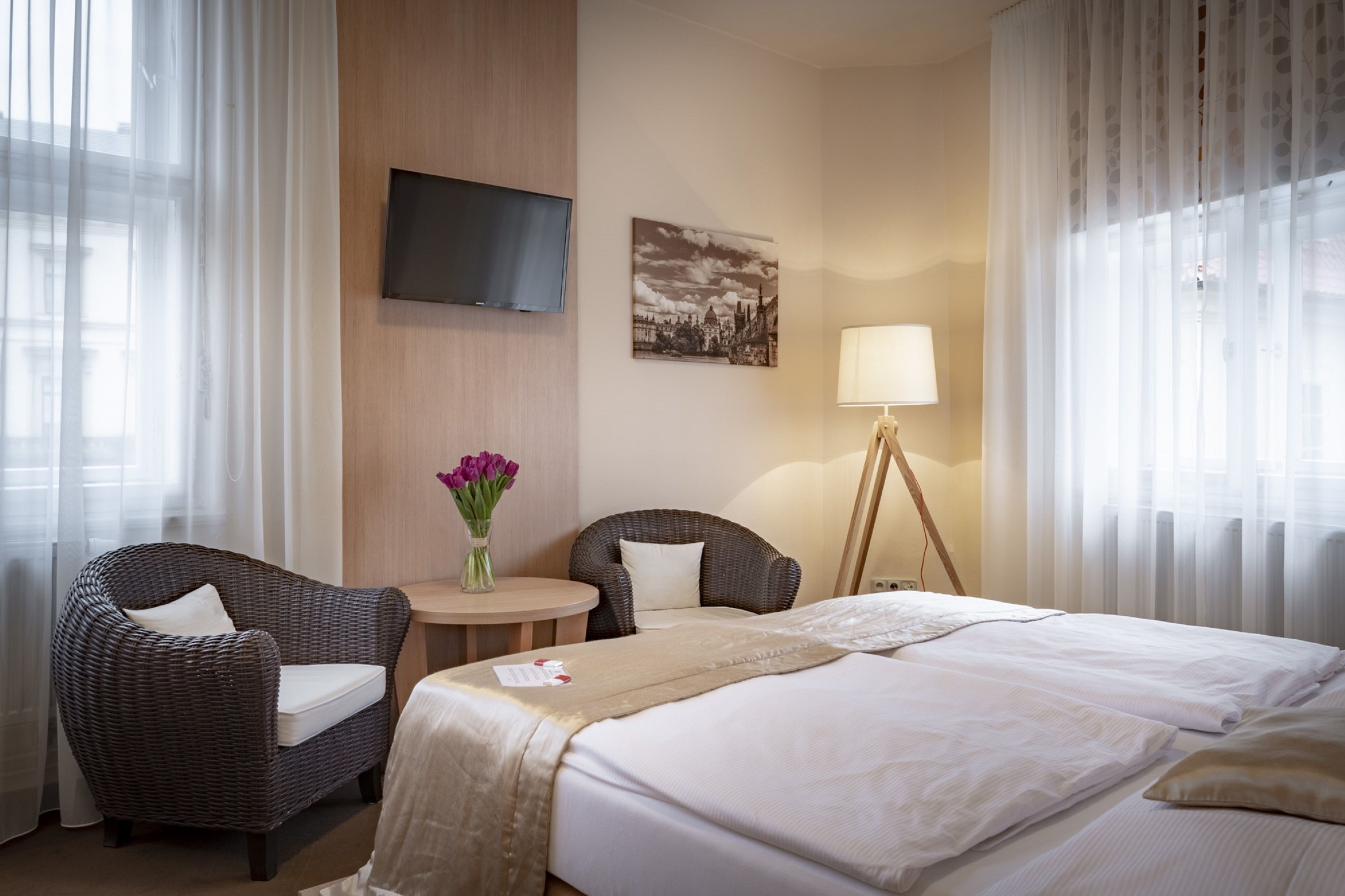 Habitació d'hotel amb un llit, dues cadires de vimetla, una taula petita amb un ram de flors, una làmpada i un televisor fixat a la paret.