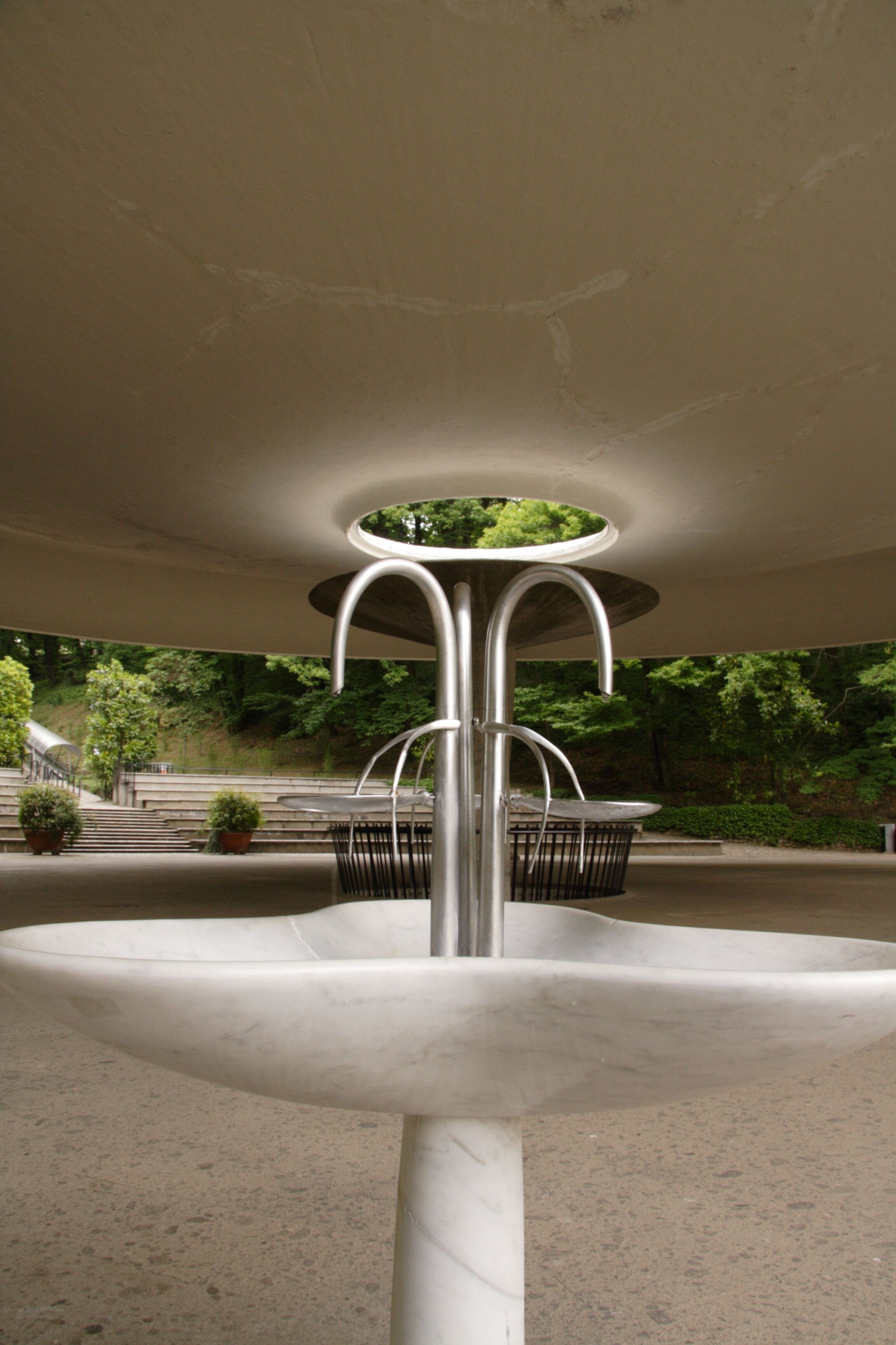 A modern outdoor fountain with a circular basin and metal supports, surrounded by a paved area and greenery.