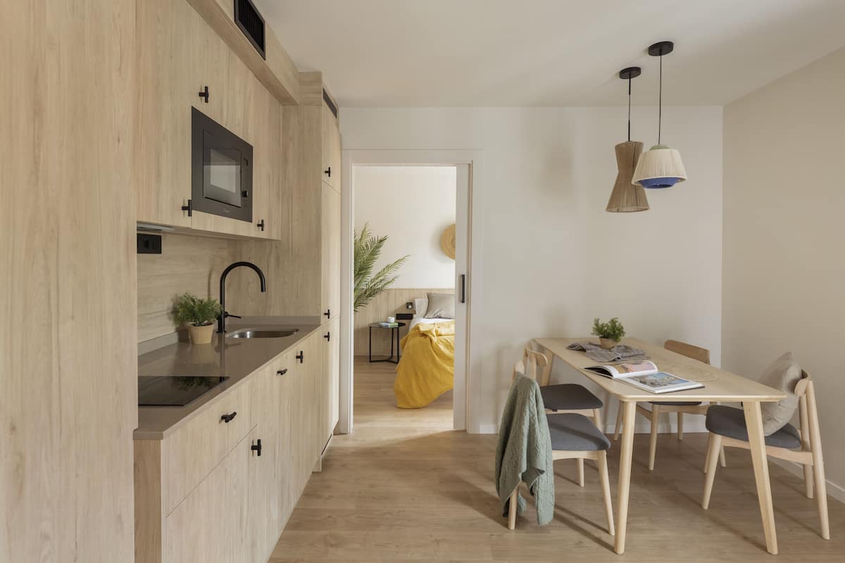 Une cuisine moderne avec des meubles en bois, un évier noir et une fenêtre. On y trouve un coin repas avec une table et des chaises, et un lit avec une couverture jaune à l’arrière-plan.