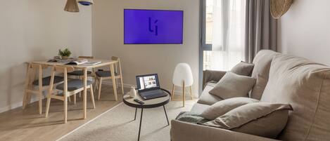 A modern living room with a sofa, a dining table with chairs, a laptop, and a flat-screen TV on the wall.