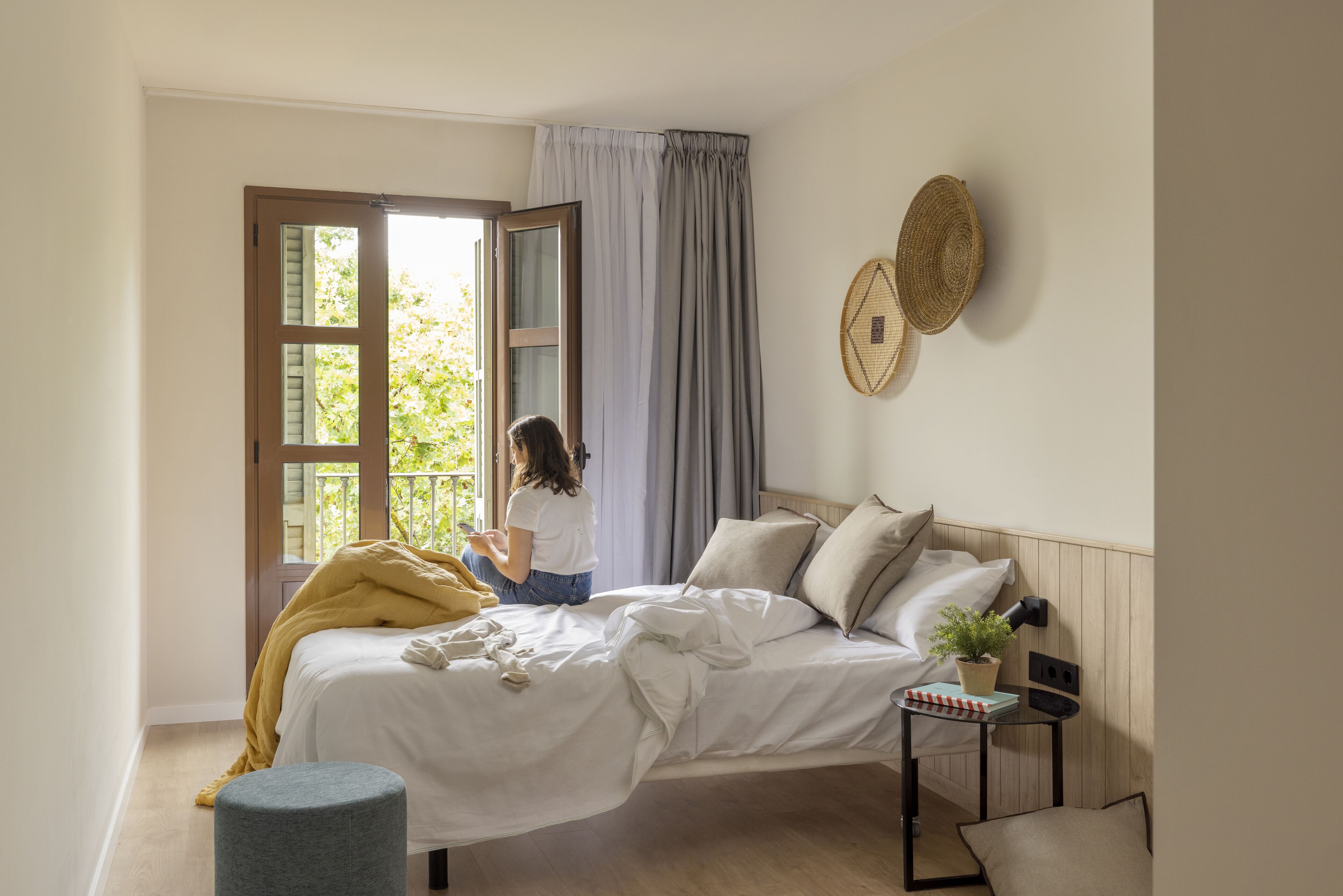 Un dormitorio con cama, mesita de noche con lámpara, ventana con cortinas y una cesta fijada a la pared.