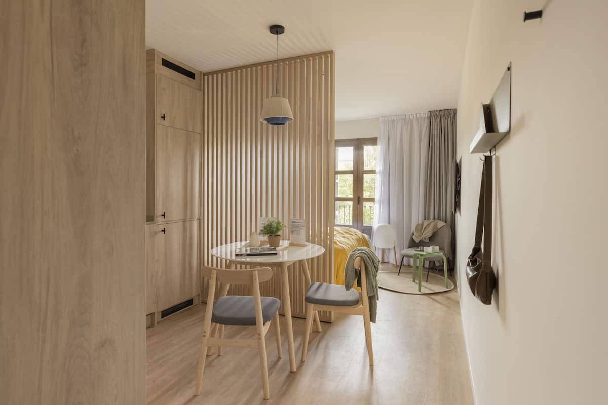 Un espace repas moderne avec une table ronde, deux chaises, un luminaire suspendu et une vue sur une pièce comprenant un lit et des rideaux.