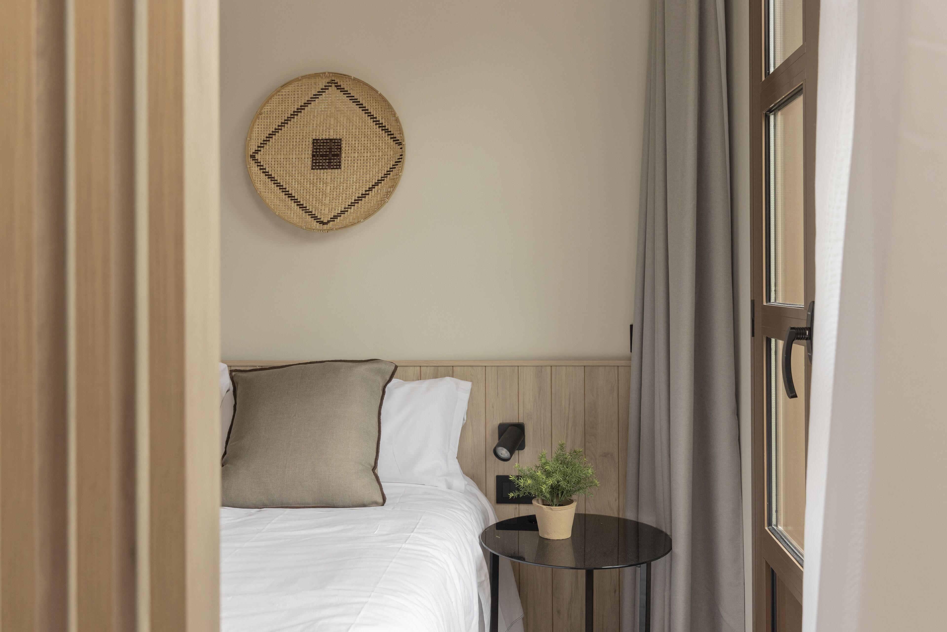 A neatly arranged hotel room with a bed, a small round table with a plant, a wall-mounted light, and a decorative woven item above the bed.