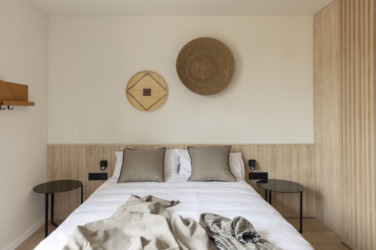 Un lit bien fait, avec une couette blanche et deux oreillers, une décoration murale ronde en fibres tressées et une petite table ronde placée à côté du lit.