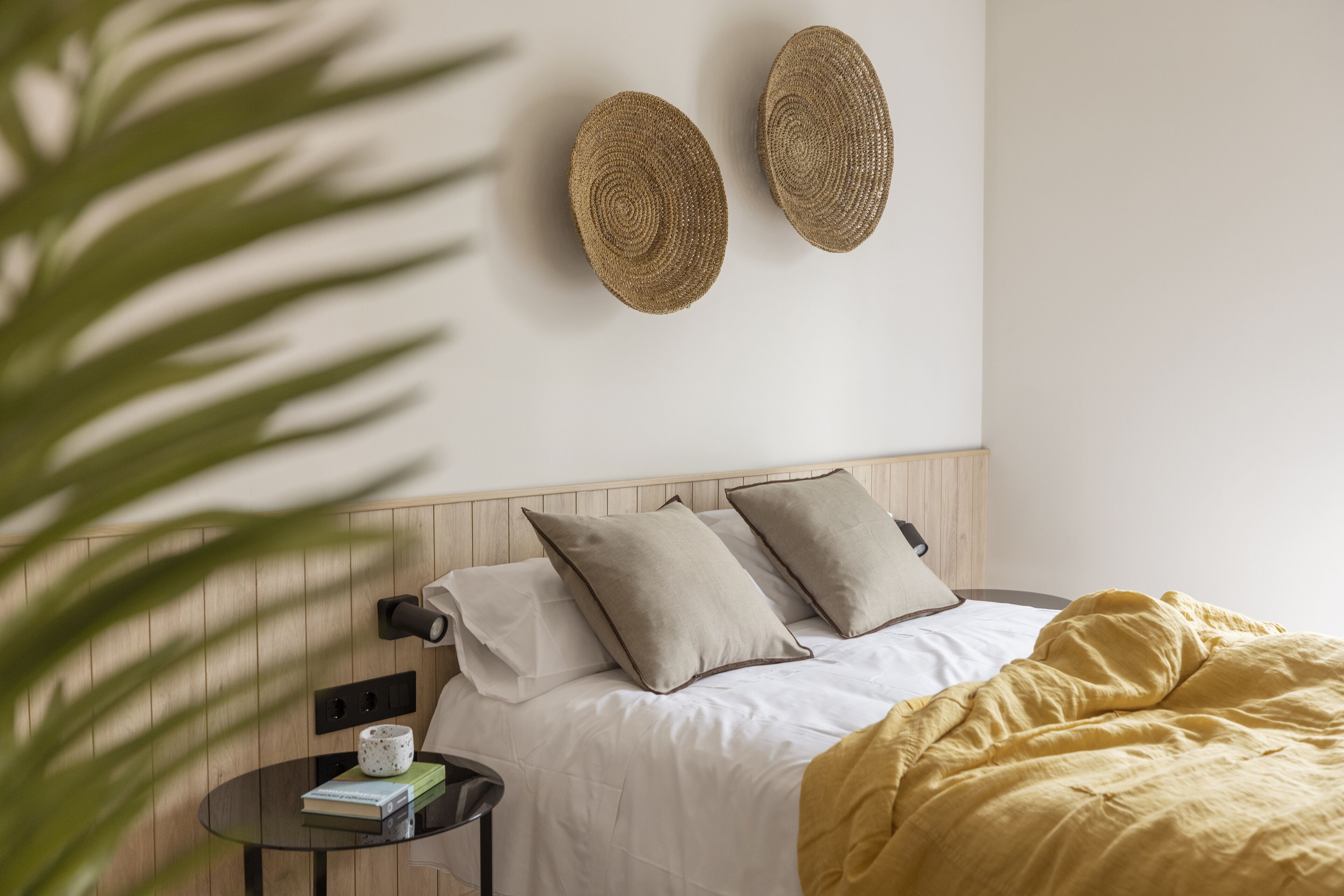 Una cama bien hecha con una manta amarilla, una mesita de noche con un libro y una taza, y dos adornos de pared tejidos.