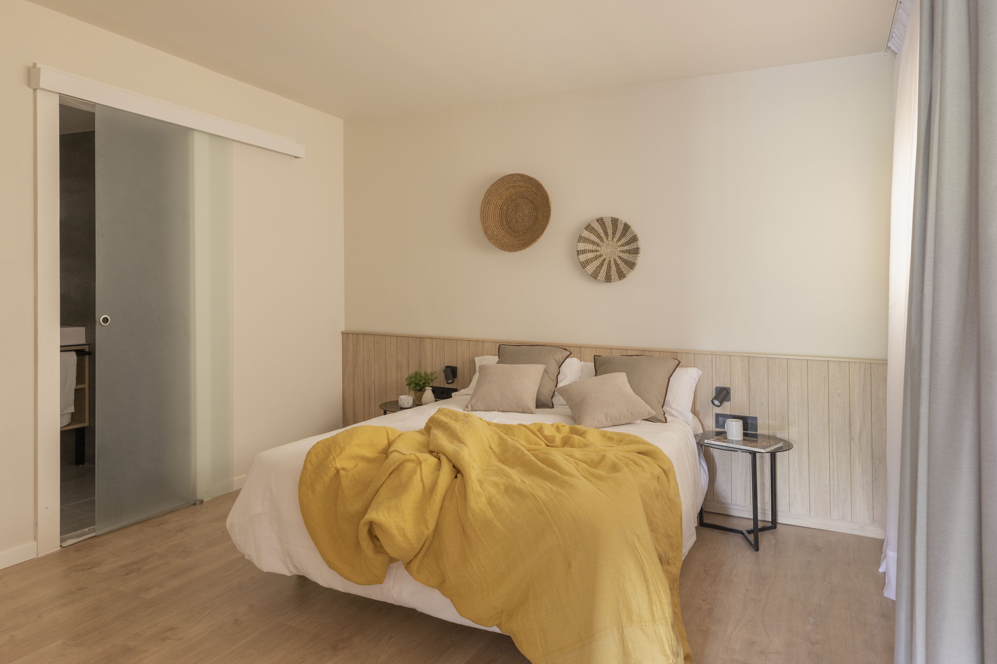 A bedroom with a bed, bedside tables, a wall-mounted clock, and a door leading to a bathroom.