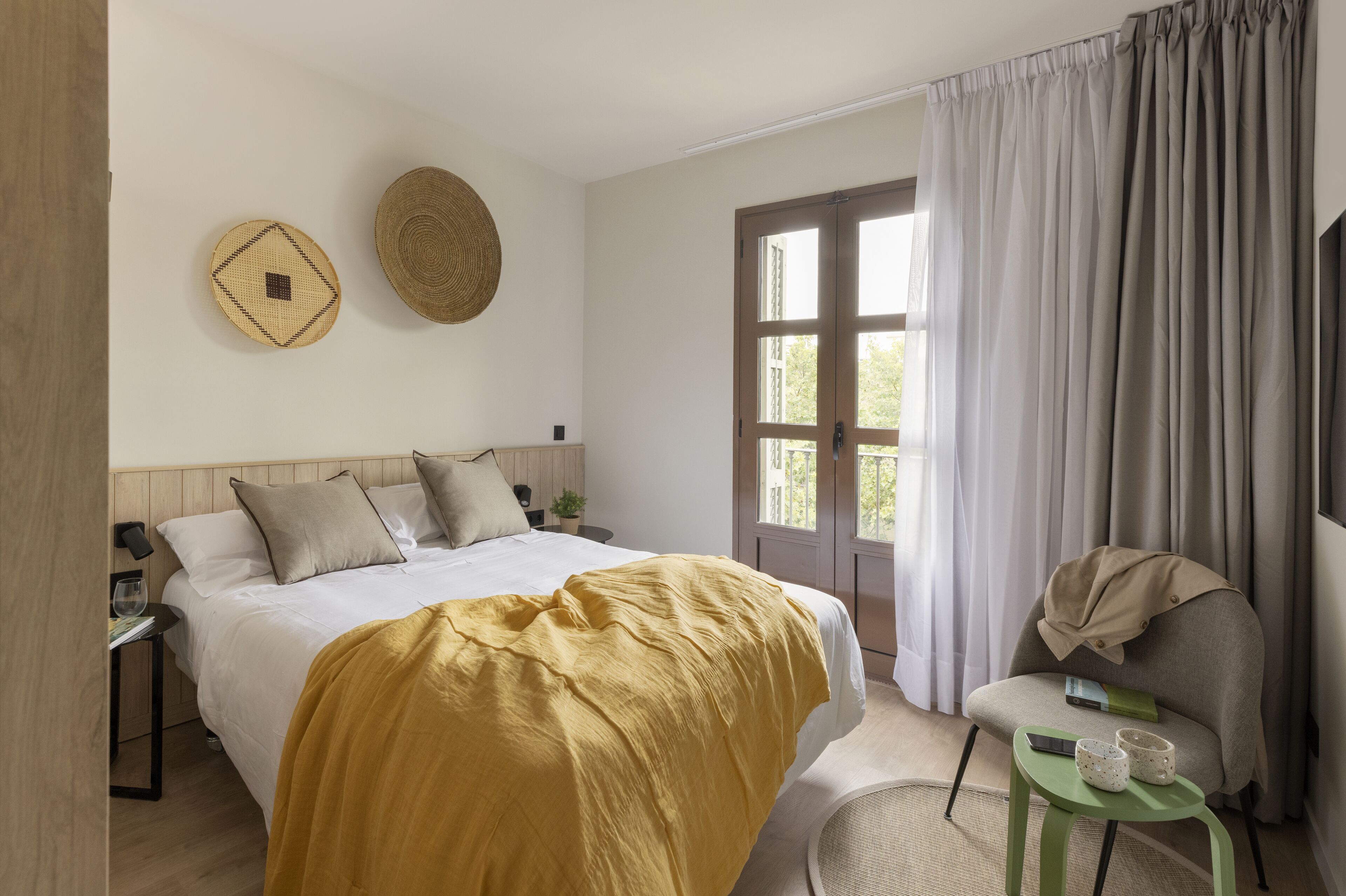 Un dormitorio ordenado con cama, una silla, una mesita y una ventana con cortinas.