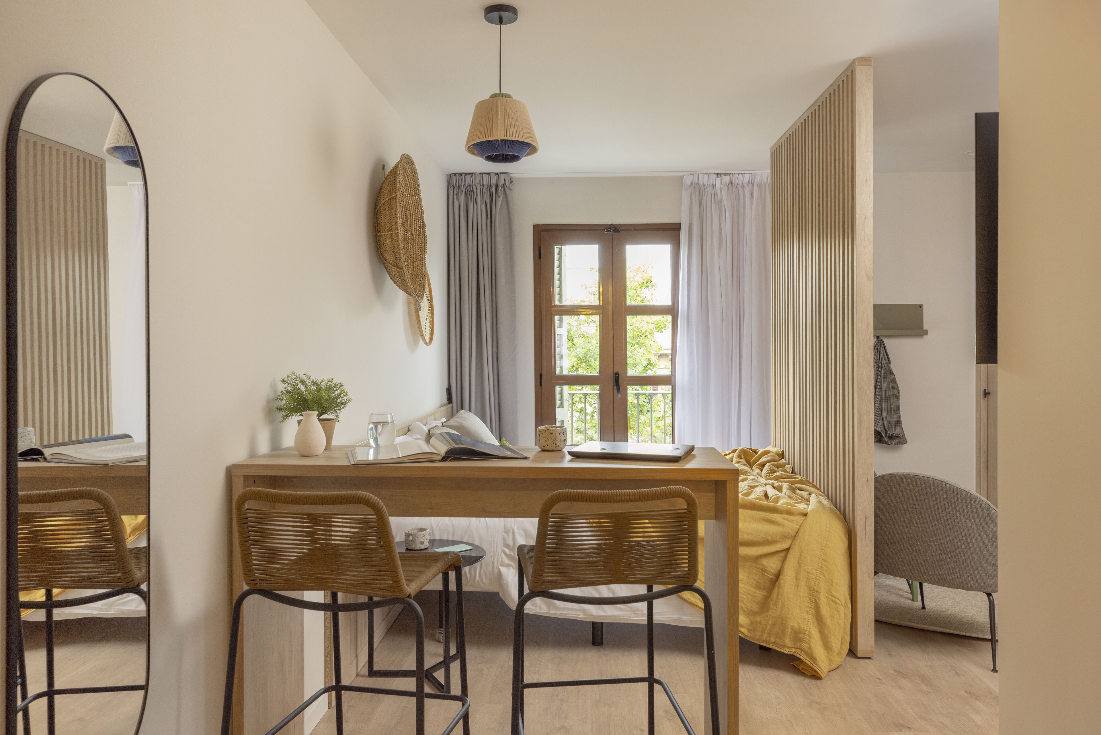 A room with a wooden desk, two wicker chairs, a bed with a yellow blanket, a large mirror, and a window with curtains.