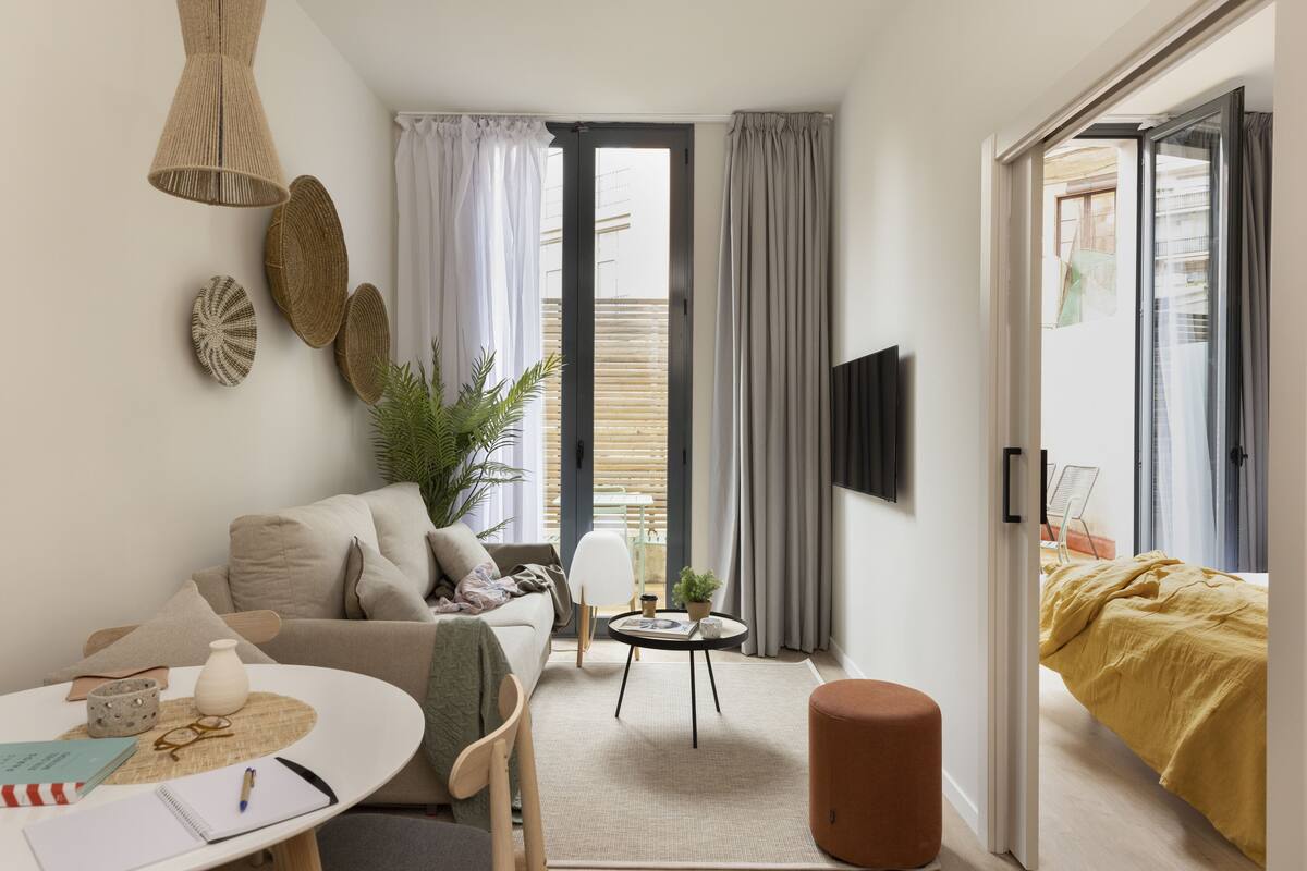 Un salon moderne et bien éclairé, avec un canapé, une petite table ronde, un téléviseur fixé au mur et une porte-fenêtre coulissante donnant sur un balcon.