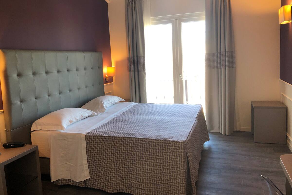 A hotel room with a large bed, a nightstand, a lamp, and a view of the outdoors through a window.