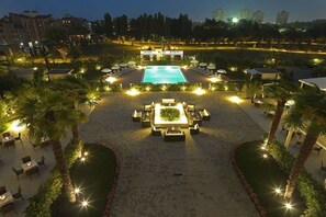 A well-lit outdoor pool area with lounge chairs and a central seating arrangement, surrounded by palm trees and a landscaped garden. - Savoia Hotel Regency (Bologna)