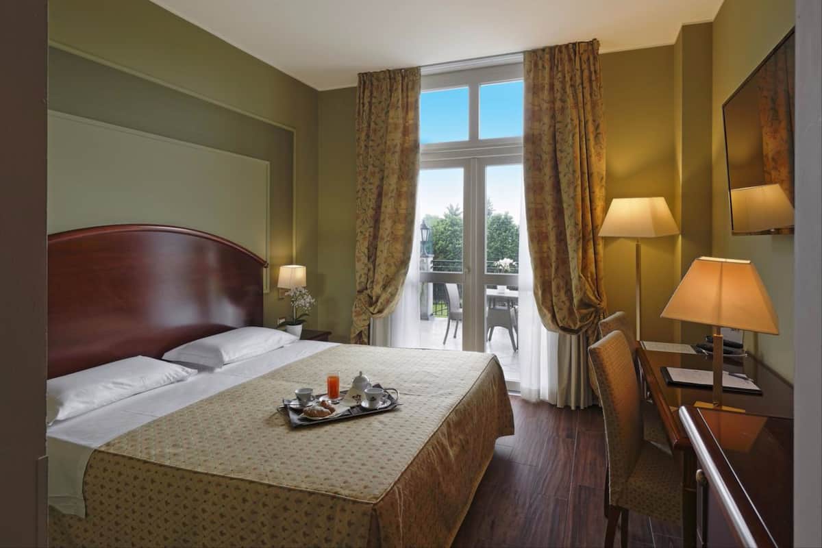 A hotel room with a bed, a desk, two lamps, a window with curtains, and a balcony view.