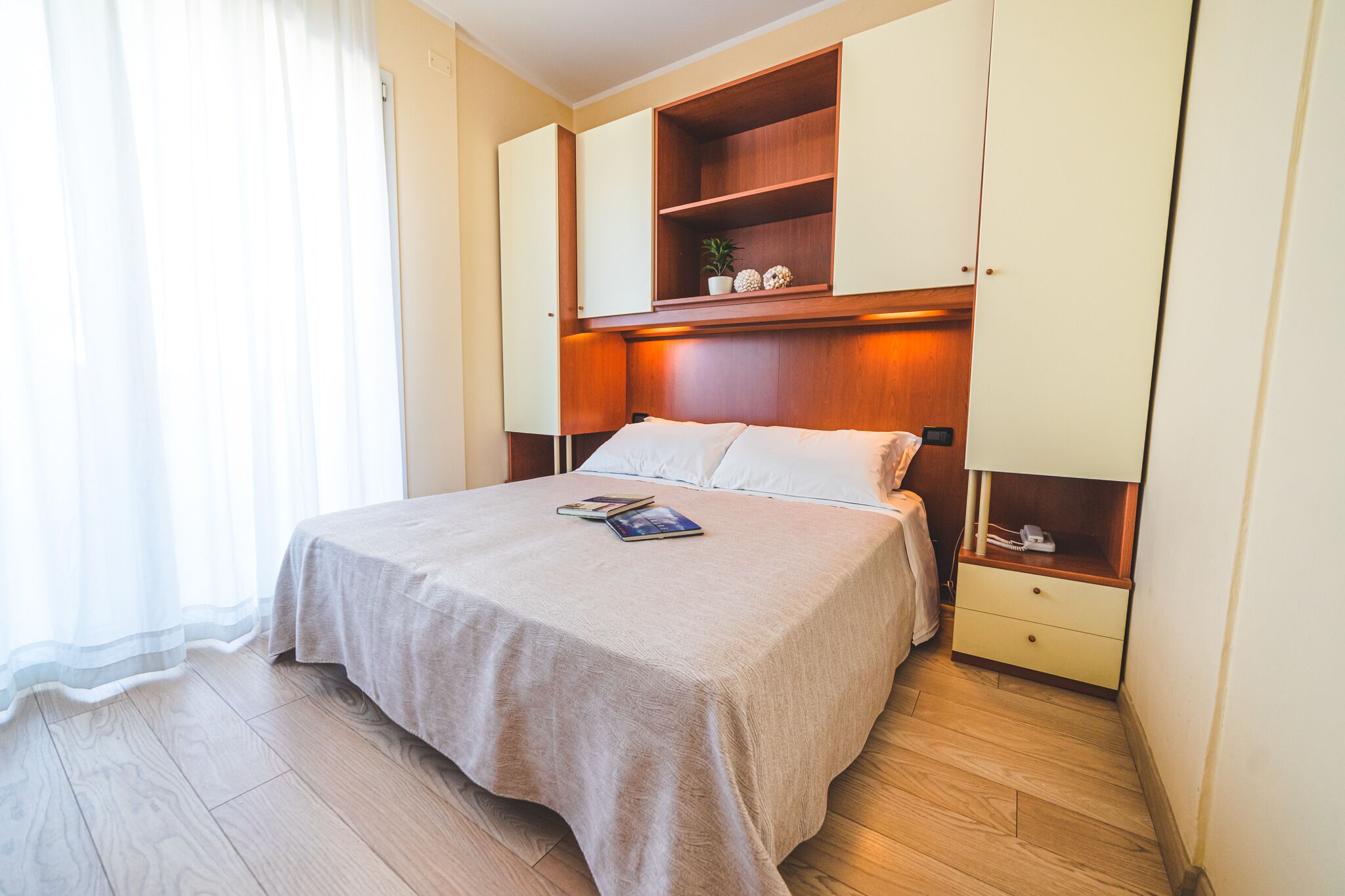 Ein Schlafzimmer mit einem Bett, einem Nachttisch, einer Holzregal mit Dekoration und einem Fenster mit durchsichtigen Vorhängen.