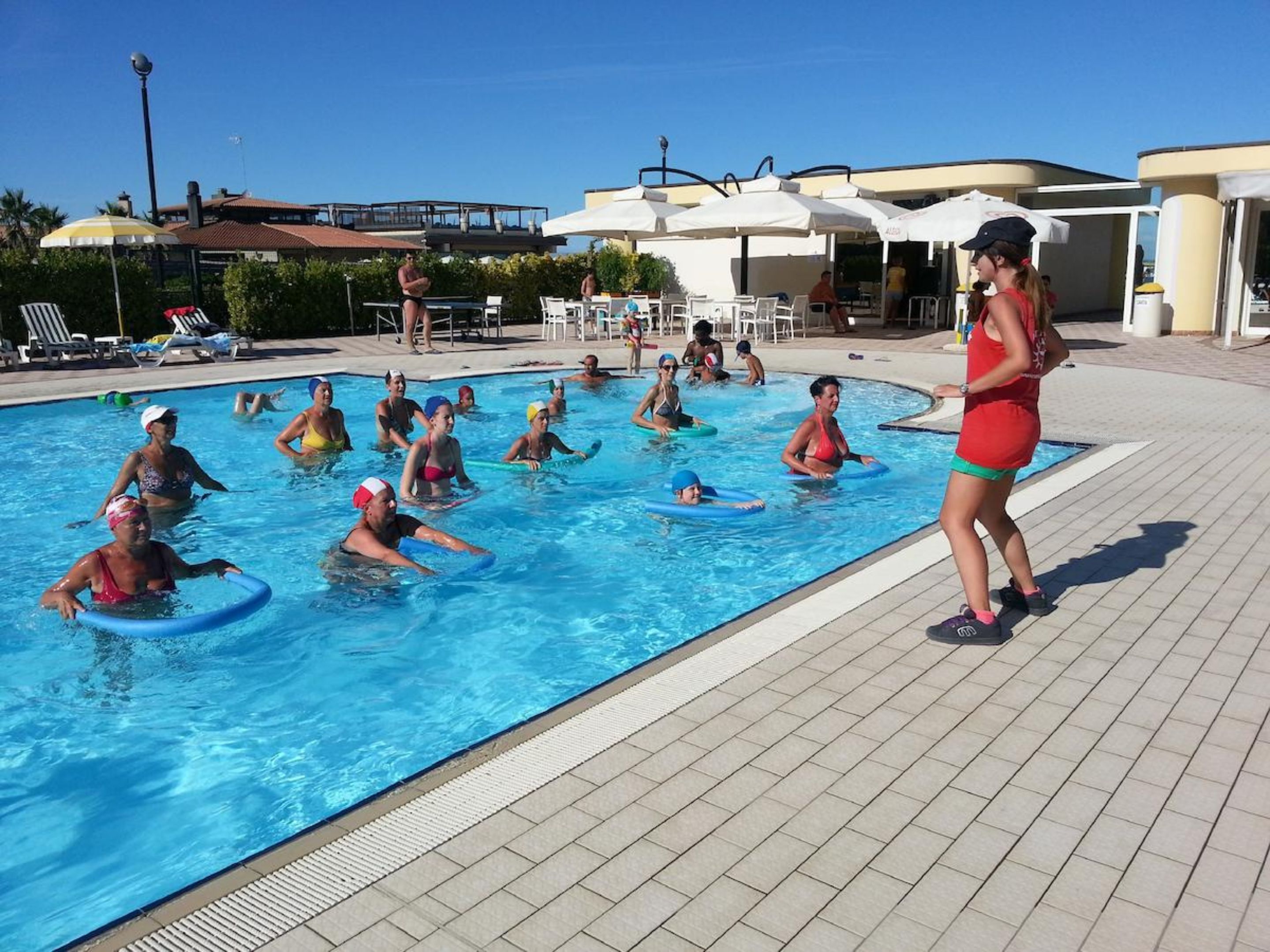 Ein Schwimmbecken mit Menschen, die Schwimmhilfen benutzen, umgeben von Liegestühlen und Sonnenschirmen.