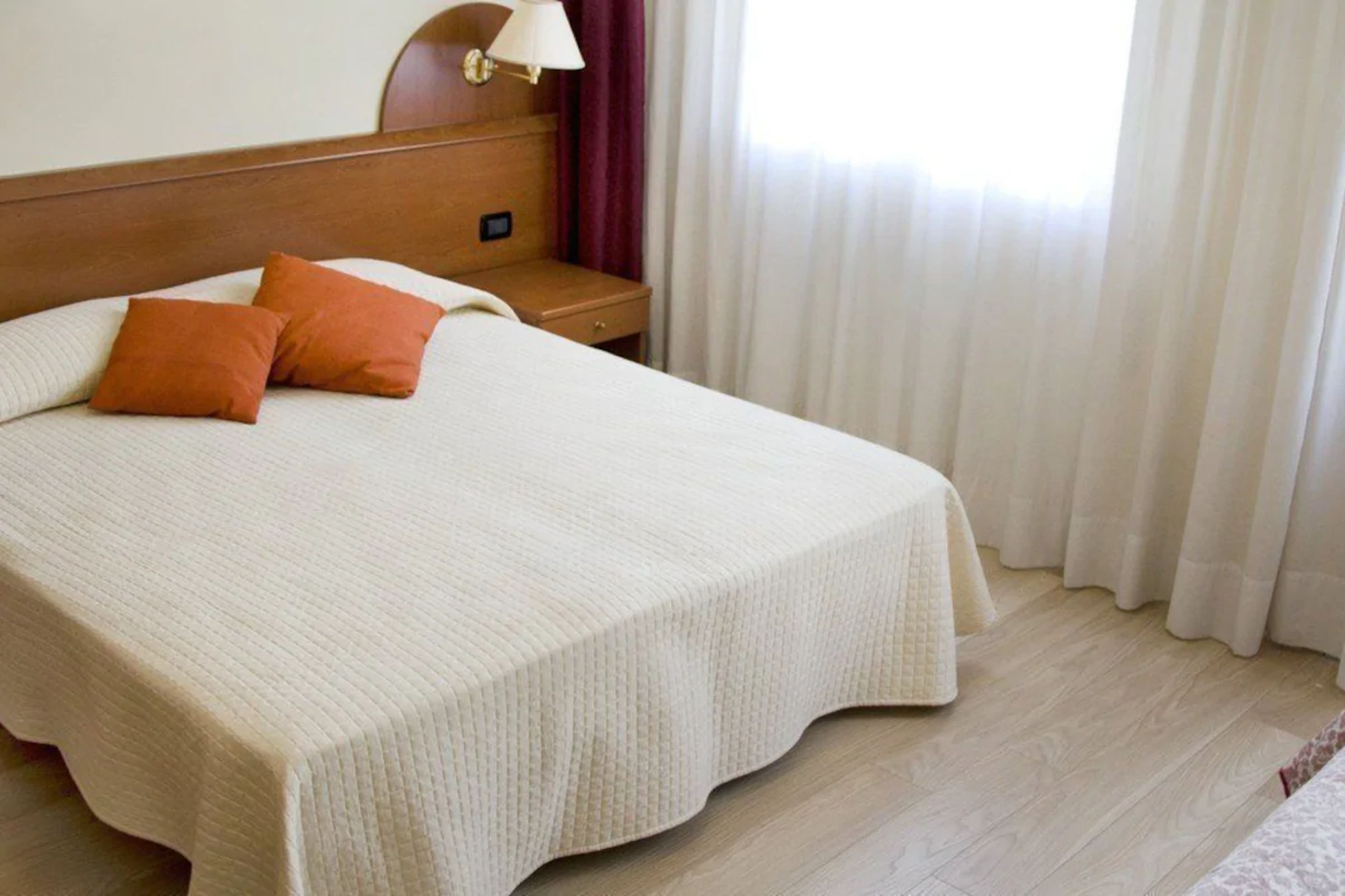Una cama bien hecha con colcha blanca y dos almohadas anaranjadas, una mesita de noche de madera con una lámpara, y una ventana con cortinas blancas.
