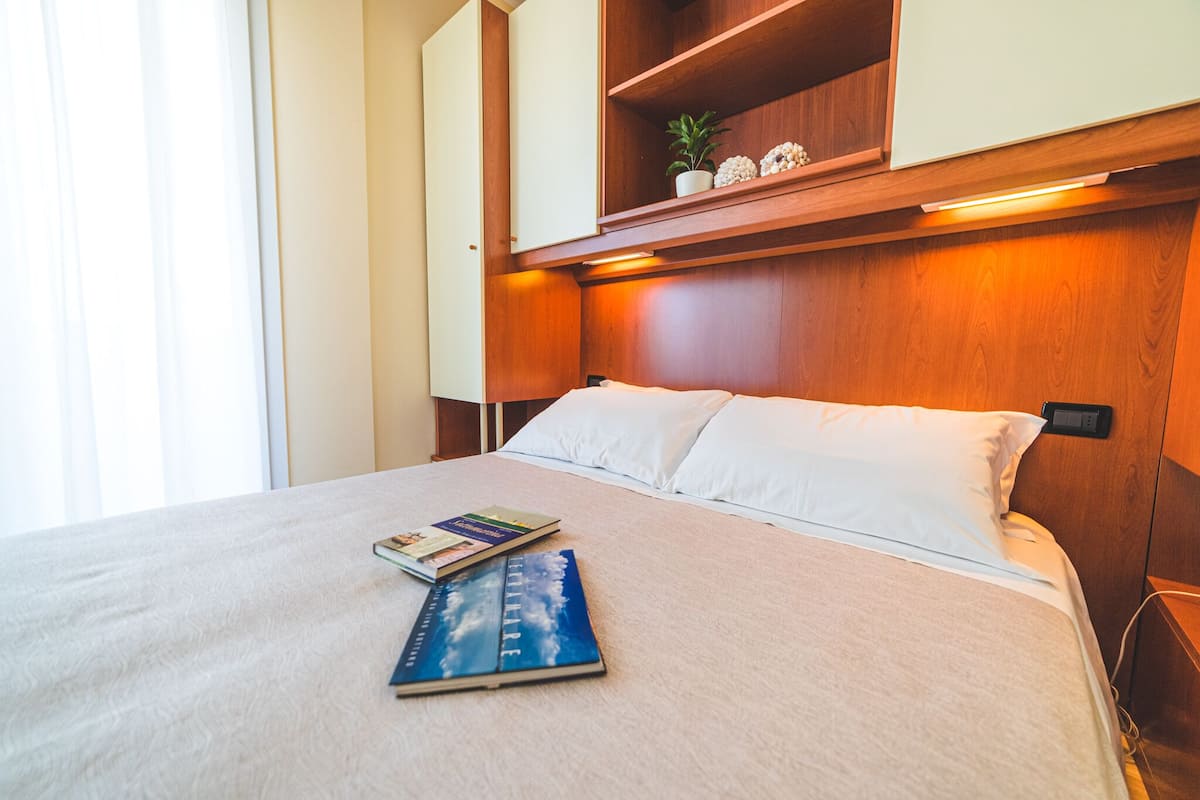 A neatly made bed with white linens and a book on it. There is a wooden shelf above the bed with decorative items.