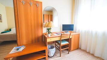 A wooden desk with a computer, a lamp, and a plant. There is a mirror on the wall and a bed in the background.