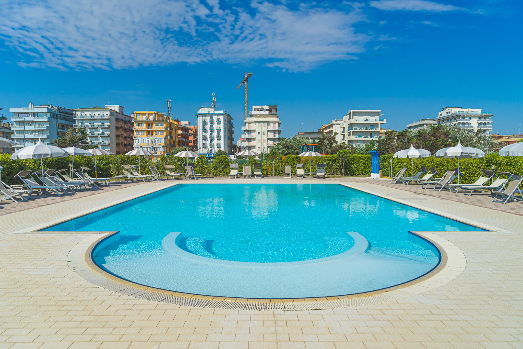 A swimming pool with lounge chairs and umbrellas, surrounded by buildings and a clear sky.