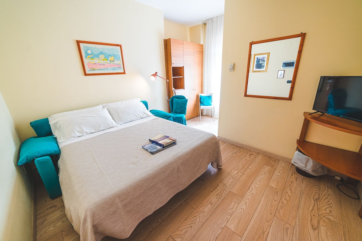 A hotel room with a bed, a television, a desk with a chair, and a wardrobe.