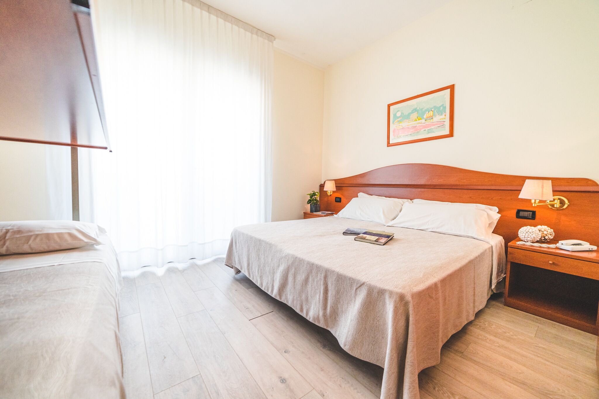 Hotelkamer met een groot bed, een kleiner bed, een nachtkastje met lamp, een schilderij aan de muur en een raam met dunne gordijnen.