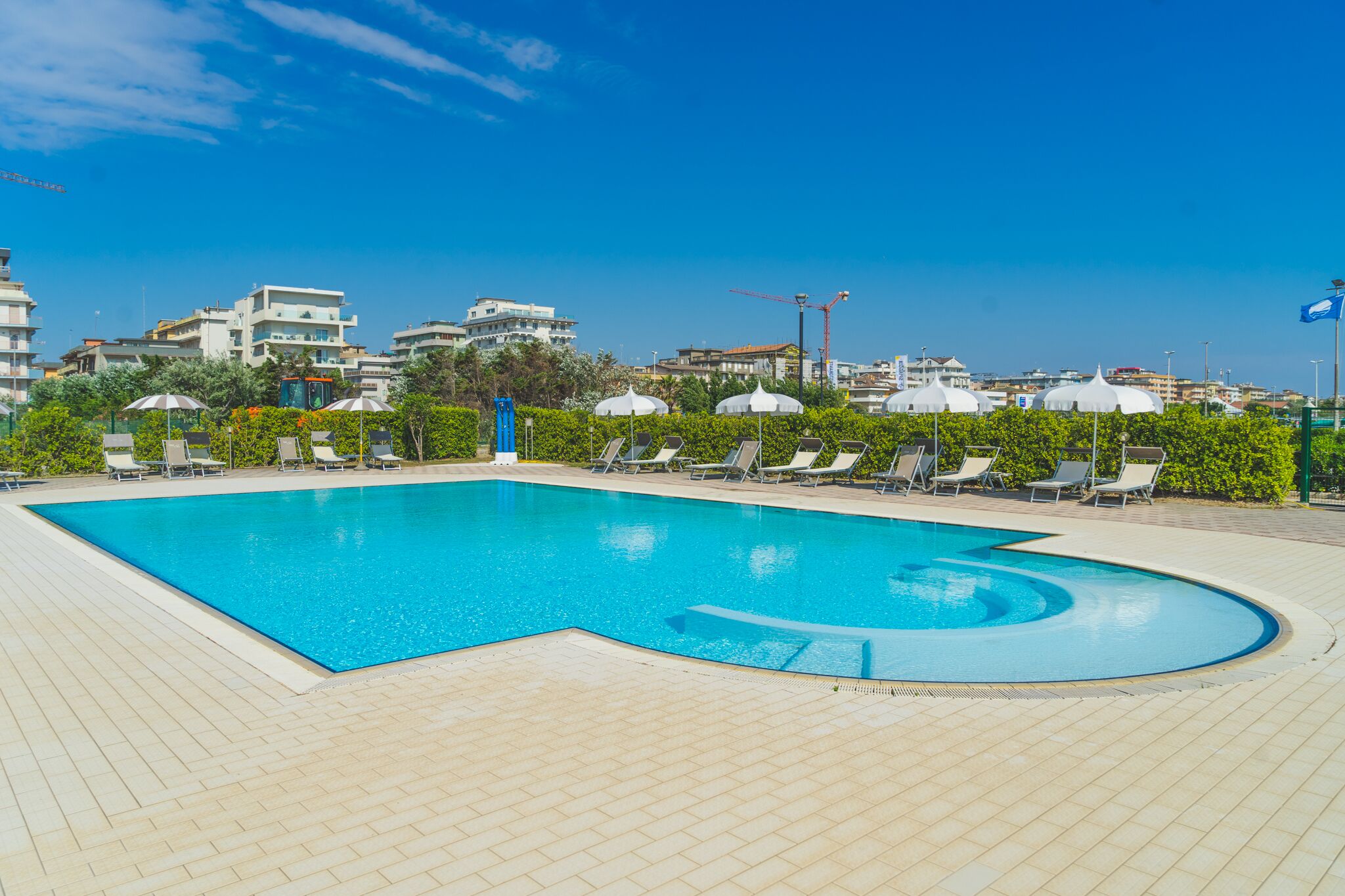 Ein Swimmingpool mit Liegestühlen und Sonnenschirmen, umgeben von Grünflächen und Gebäuden im Hintergrund.