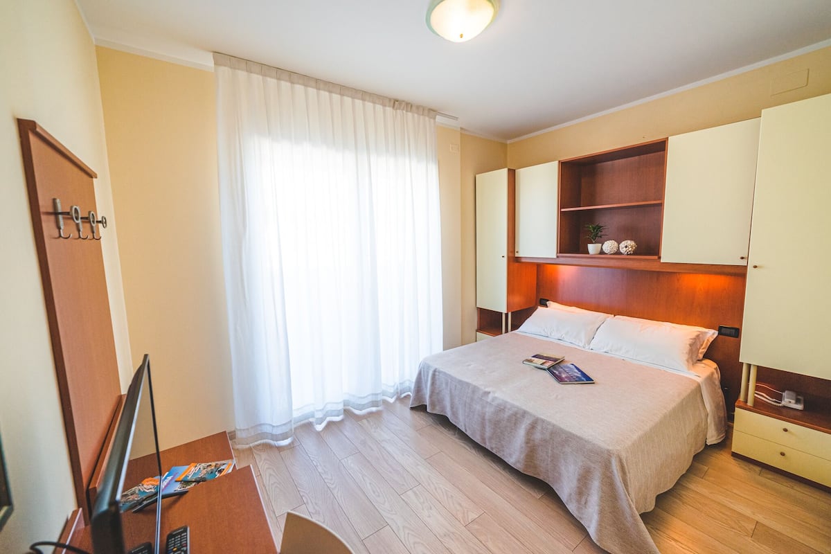 A hotel room with a bed, a desk with a computer, a TV, a wardrobe, and a window with curtains.