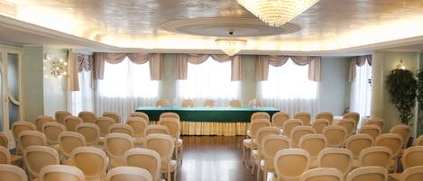 Un salón de conferencias con filas de sillas mirando a una mesa, una araña y ventanas grandes con cortinas.