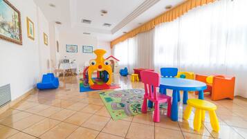 Une pièce lumineuse avec un espace de jeux comprenant une structure colorée, une table avec des chaises et un tapis orné d’un motif représentant un paysage urbain.