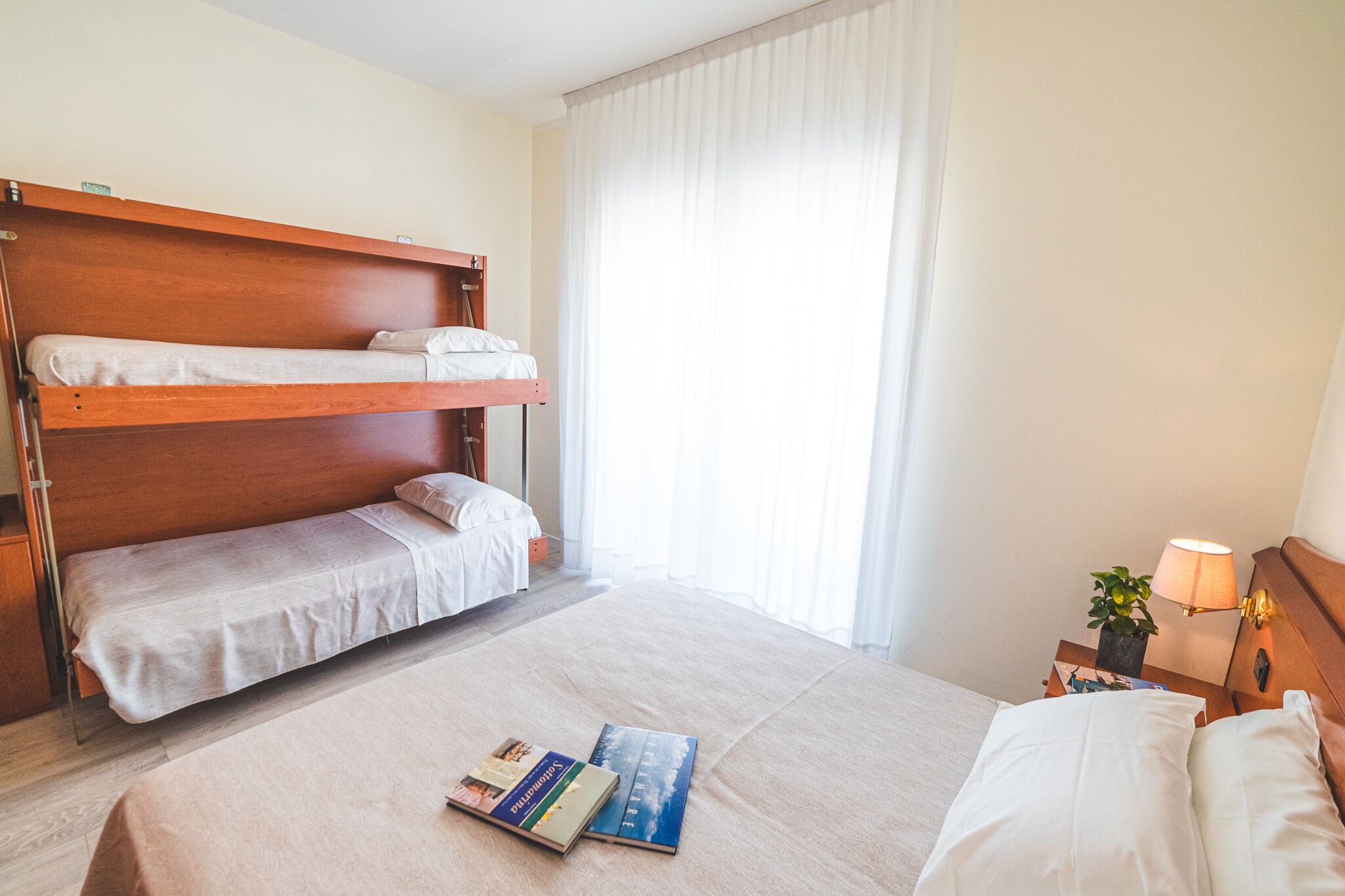 A hotel room with a bunk bed, a single bed, a nightstand with a lamp, and a window with curtains.