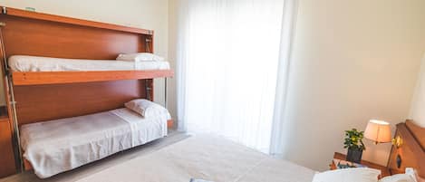 A hotel room with a bunk bed, a single bed, a nightstand with a lamp, and a window with curtains.