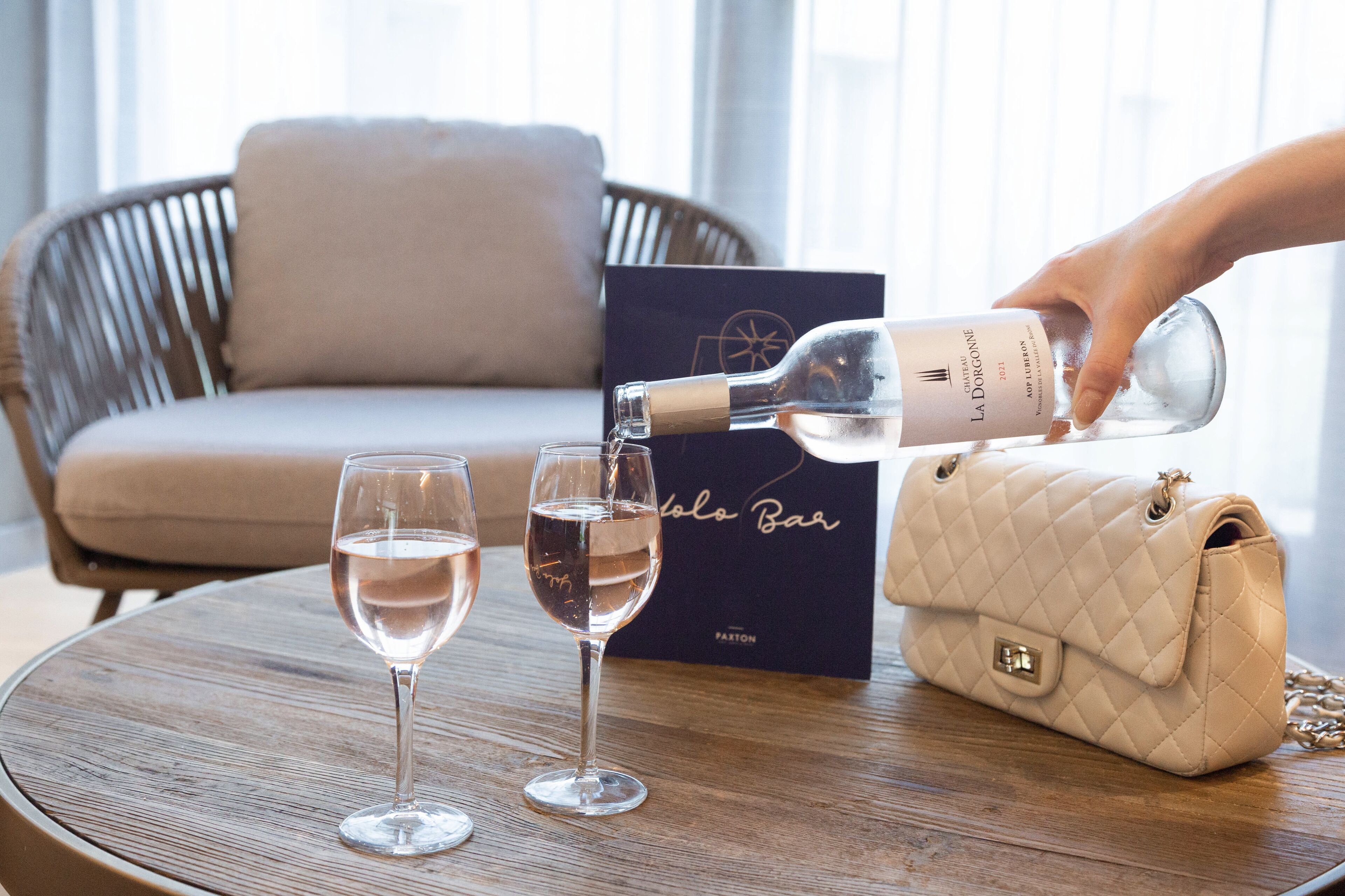 Eine Flasche La Dorconne Wein wird in zwei Gläser auf einem Holztisch gegossen, im Hintergrund eine beige gesteppte Handtasche und eine dunkelblaue Schachtel mit goldener Schrift.