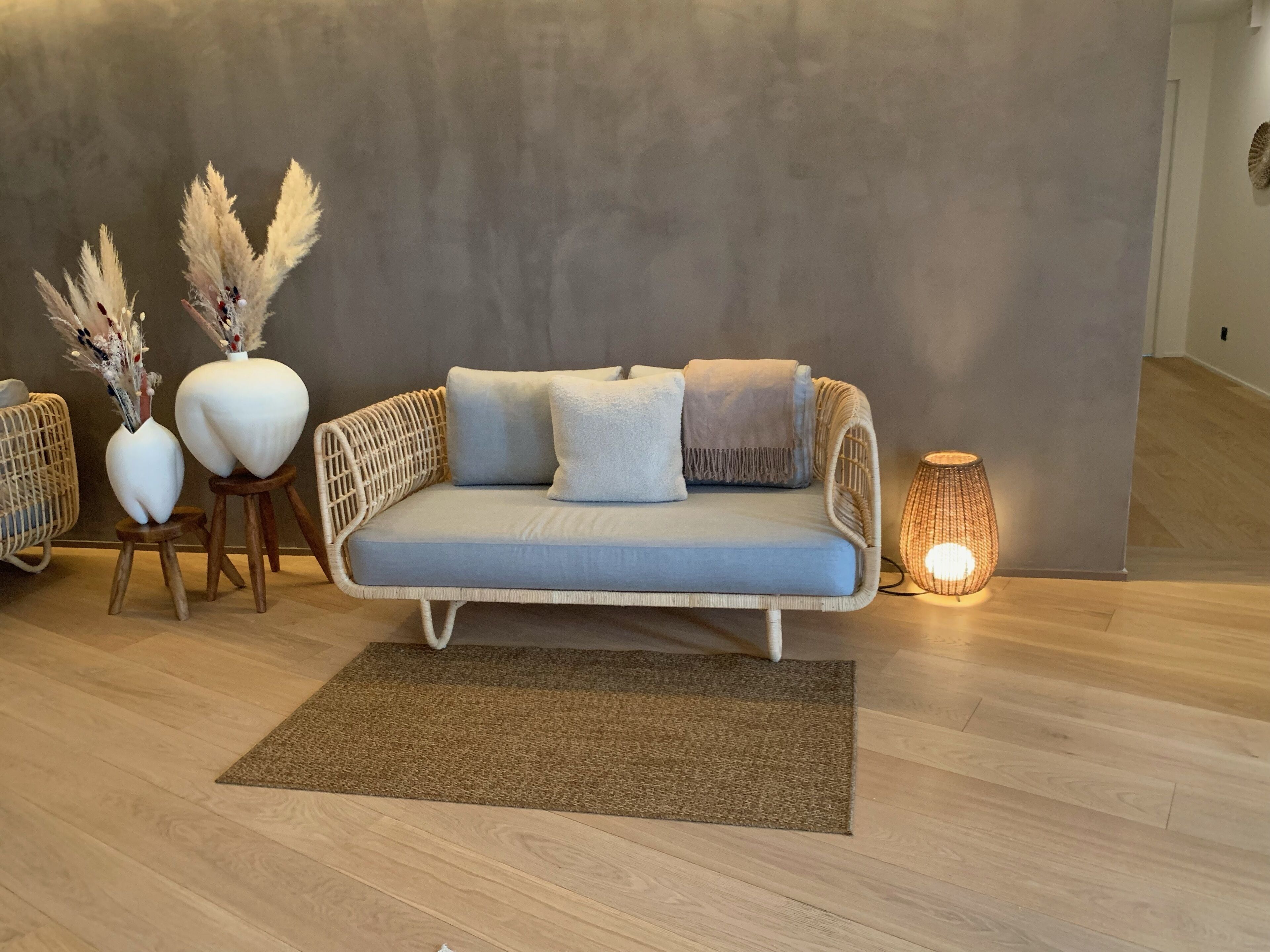 A modern living room with a light blue sofa, two white vases with pampas grass, a woven side table, a brown rug, and a lit floor lamp.