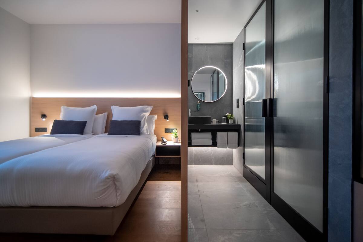 A modern hotel room with a large bed, bedside tables, a nightstand, a bathroom with a sink and mirror, and a walk-in closet.